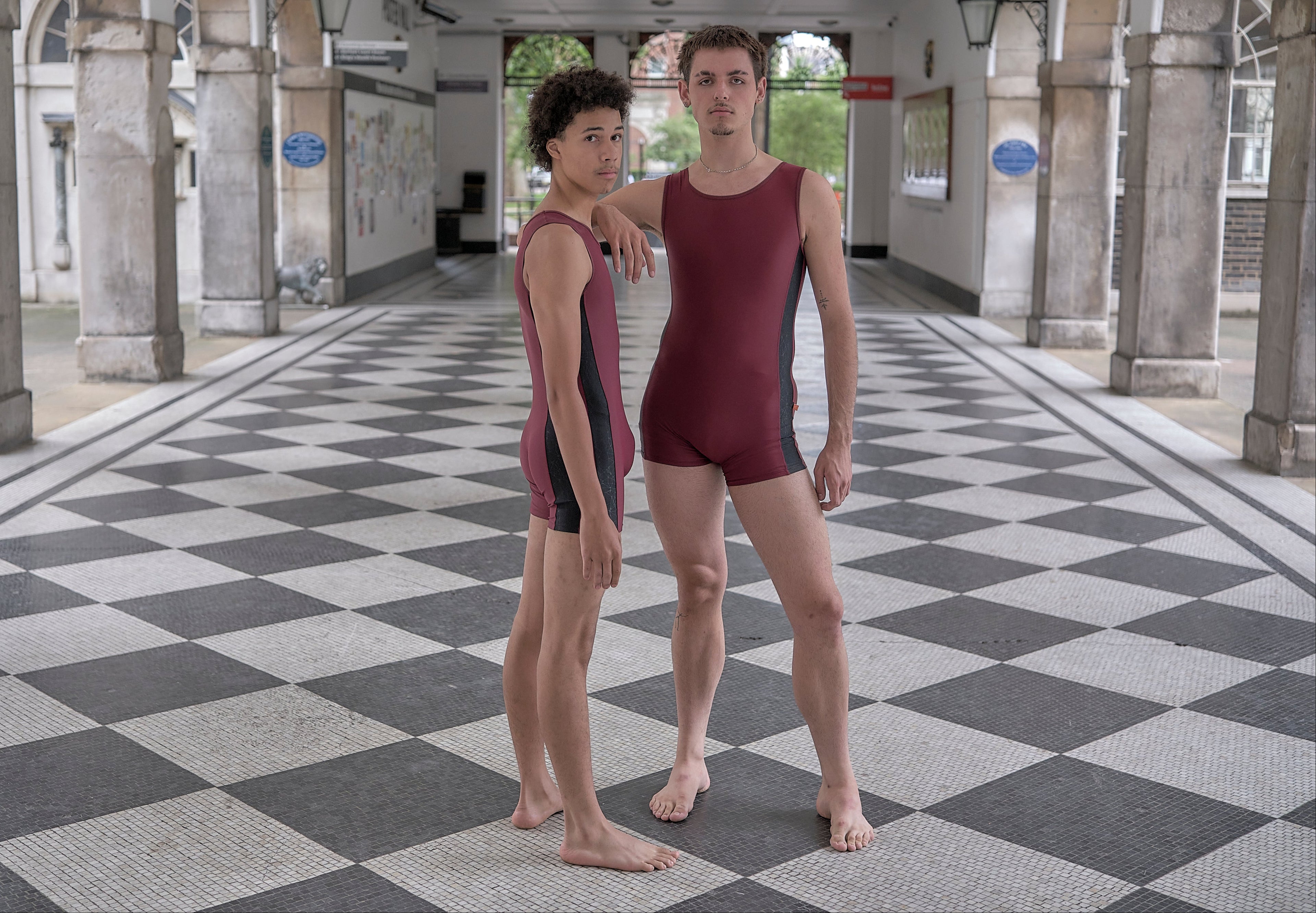 Two male dancers in maroon unitards standing on a checkered floor with columns in the background.